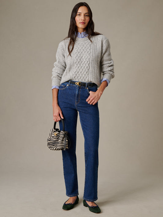 Woman wearing J.McLaughlin Willow Jeans paired with a gray cable-knit sweater, blue collared shirt, black belt, green flats, and holding a zebra-print bag, showcasing timeless style.