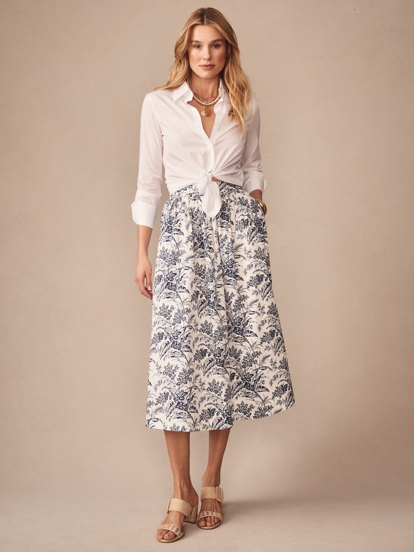 A model wearing the Winnie Skirt in Giardino Toile by J.McLaughlin, featuring a white and navy floral pattern, styled with a tied white blouse and neutral sandals, exemplifying effortlessly stylish, enduring designs and the legacy of timeless fashion in a destination for defining style. Life, Well-Lived.