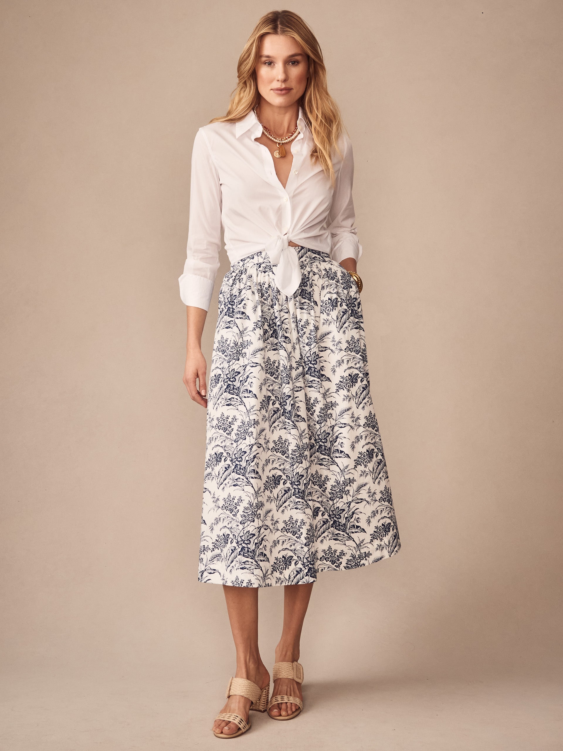 A model wearing the Winnie Skirt in Giardino Toile by J.McLaughlin, featuring a white and navy floral pattern, styled with a tied white blouse and neutral sandals, exemplifying effortlessly stylish, enduring designs and the legacy of timeless fashion in a destination for defining style. Life, Well-Lived.