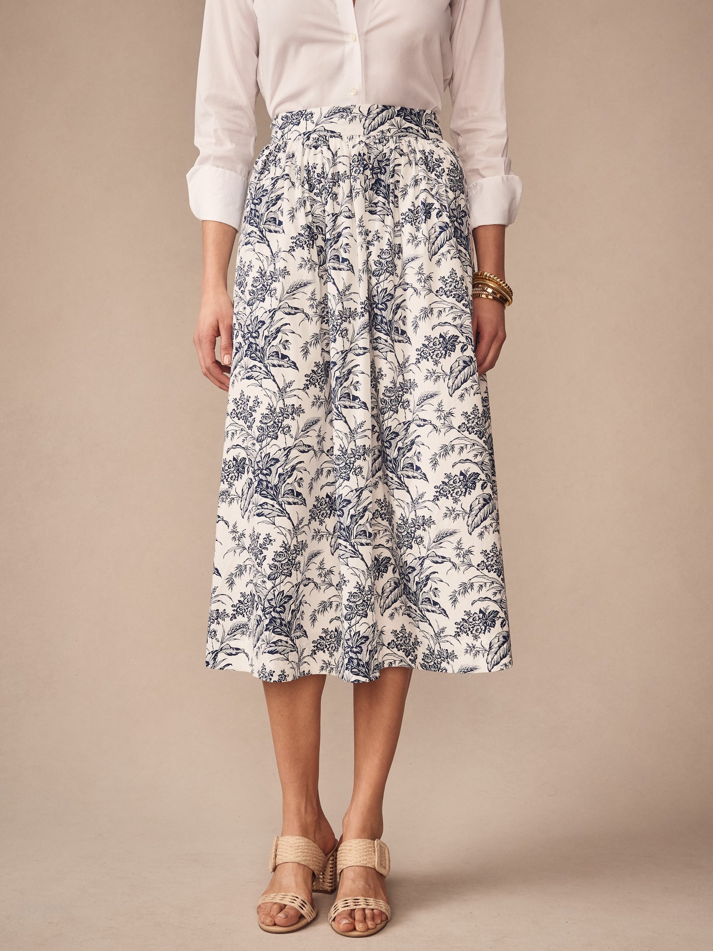 A model wearing the Winnie Skirt in Giardino Toile by J.McLaughlin, featuring a white and navy floral pattern paired with a classic white blouse and neutral sandals, showcasing effortlessly stylish skirts with enduring designs crafted from innovative fabrics. A destination for defining style. Life, Well-Lived.