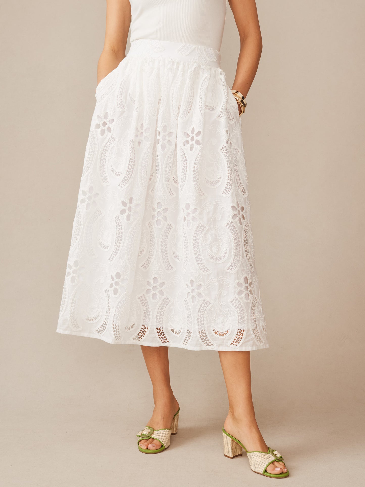 Woman wearing a white Winnie Skirt in Eyelet by J.McLaughlin with intricate floral cutout details, paired with a simple top and stylish green heels, showcasing effortlessly stylish, enduring designs and a legacy of timeless fashion. A destination for defining style. Life, Well-Lived.