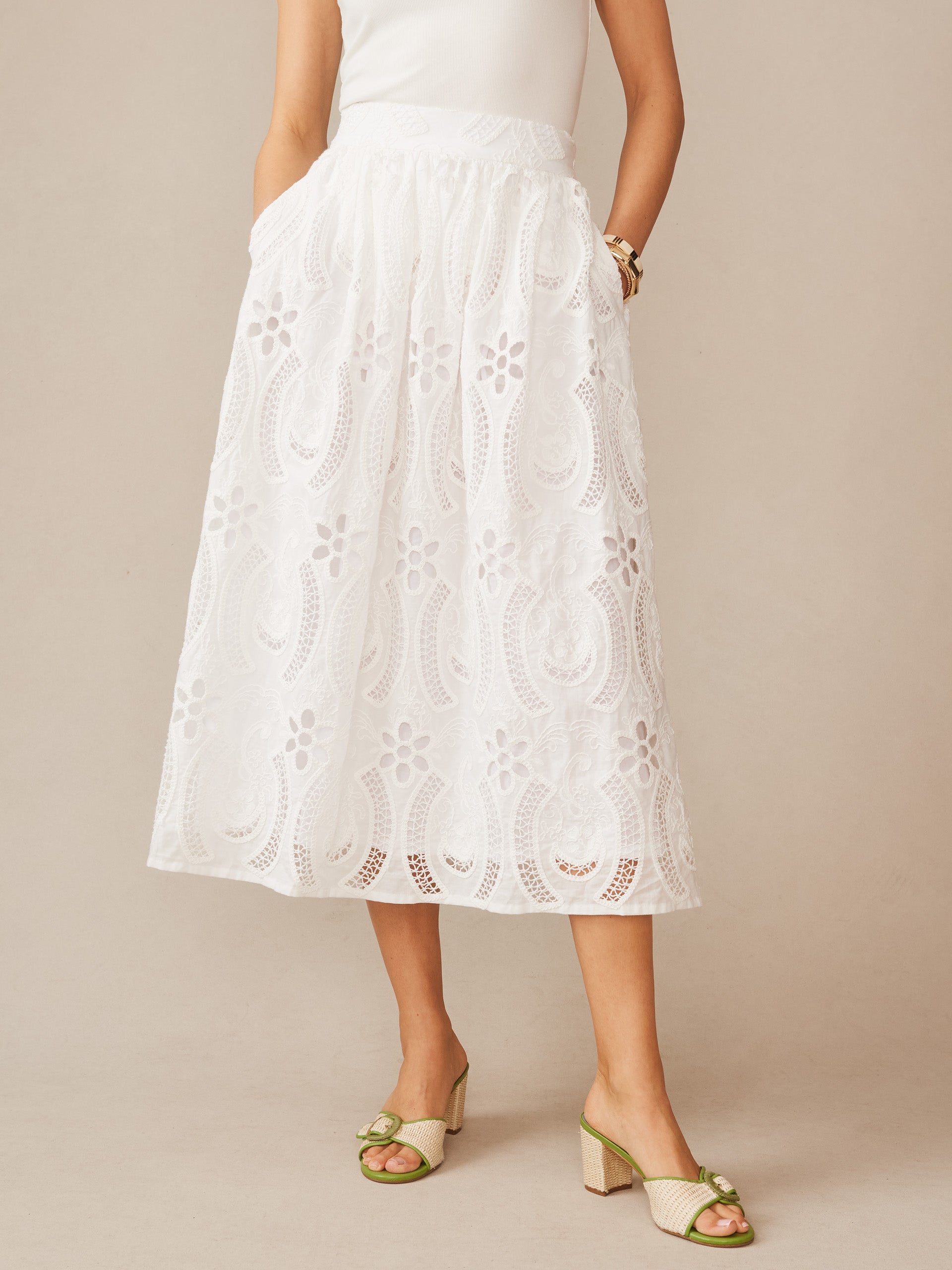 Woman wearing a white Winnie Skirt in Eyelet by J.McLaughlin with intricate floral cutout details, paired with a simple top and stylish green heels, showcasing effortlessly stylish, enduring designs and a legacy of timeless fashion. A destination for defining style. Life, Well-Lived.
