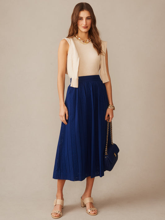 Woman wearing a sleeveless beige top with a cream sweater draped over her shoulders and a navy blue Winnie Skirt in Eyelet by J.McLaughlin, paired with neutral sandals and a navy handbag, showcasing effortlessly stylish, enduring designs and innovative fabrics from a destination for defining style. Life, Well-Lived.