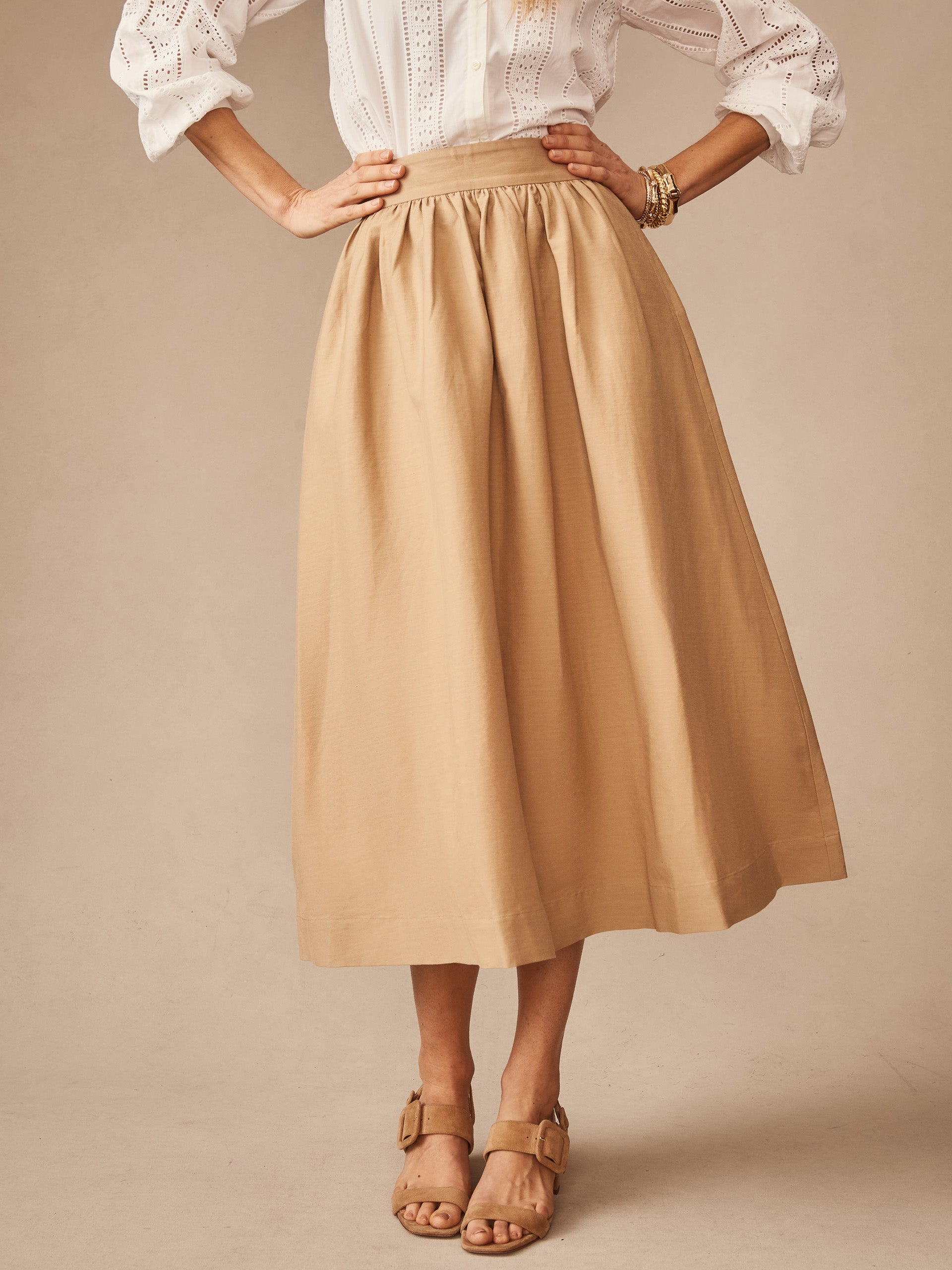 A woman wearing a khaki Winnie Linen Blend Skirt by J.McLaughlin, paired with a white eyelet blouse and tan sandals, showcasing effortlessly stylish and enduring designs.