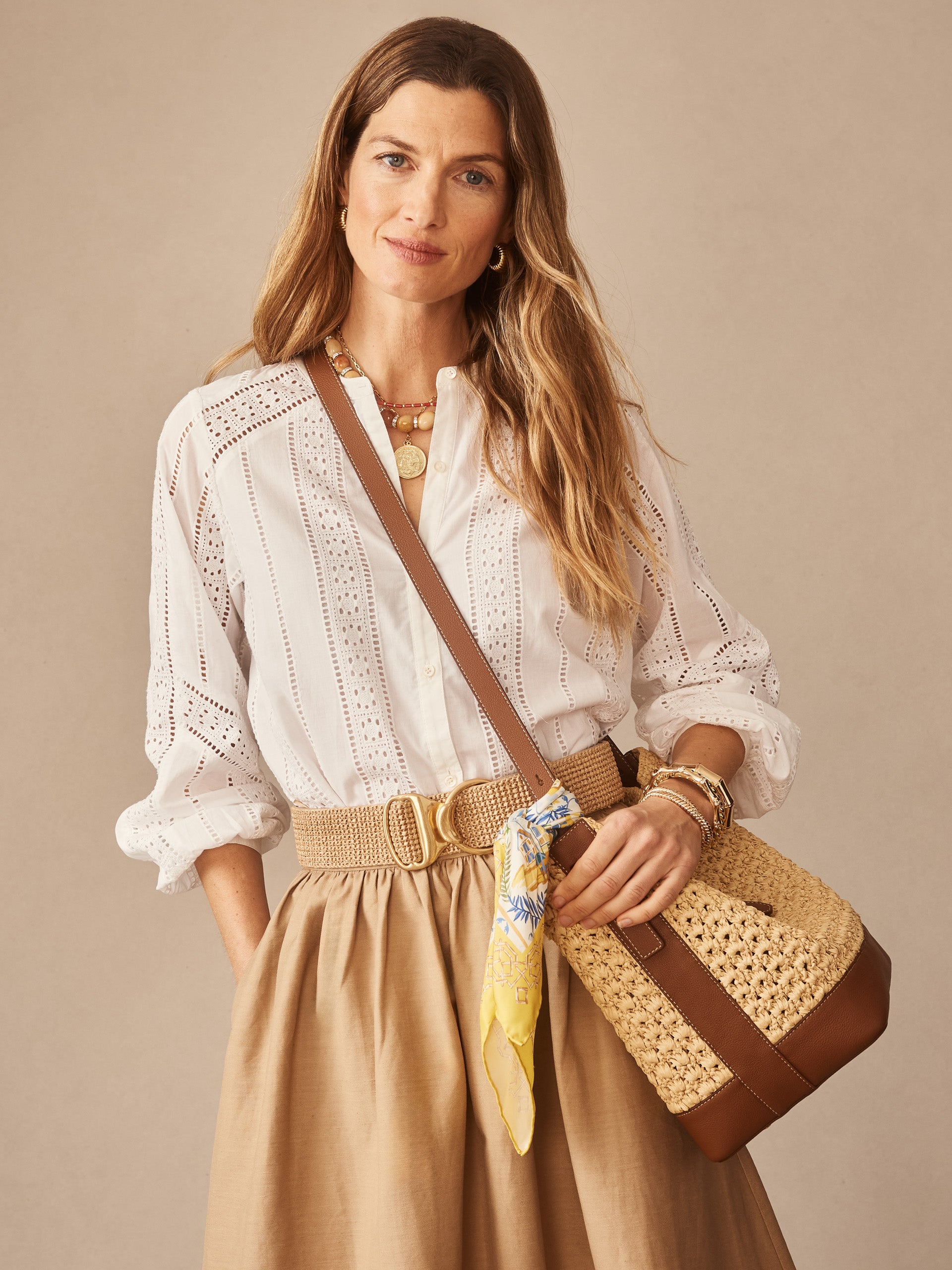 Woman wearing a white Hazel Blouse in Eyelet by J.McLaughlin paired with a high-waisted beige skirt, accessorized with a woven belt and a straw bag with leather accents and a yellow scarf, embodying effortlessly stylish, enduring designs and a legacy of timeless fashion at a destination for defining style. Life, Well-Lived.