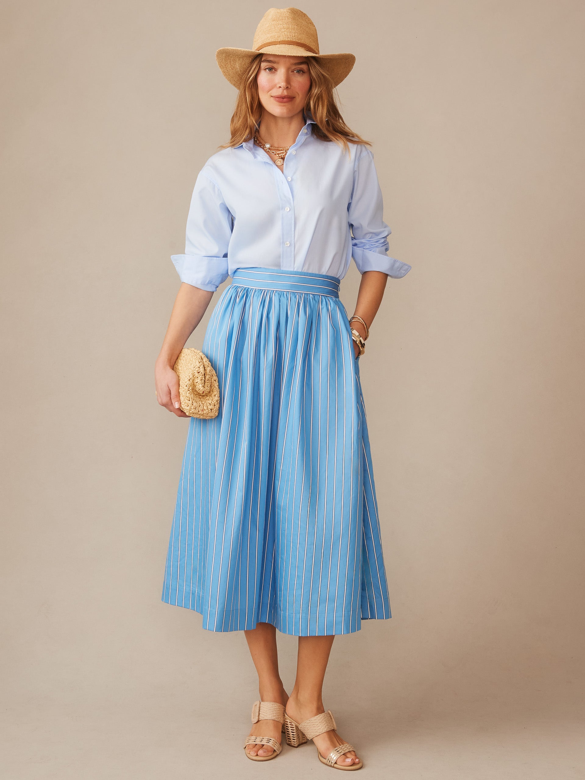 Woman wearing a Winnie Skirt in Stripe by J.McLaughlin in blue with white stripes, paired with a light blue shirt, straw hat and clutch, showcasing effortlessly stylish, enduring designs.