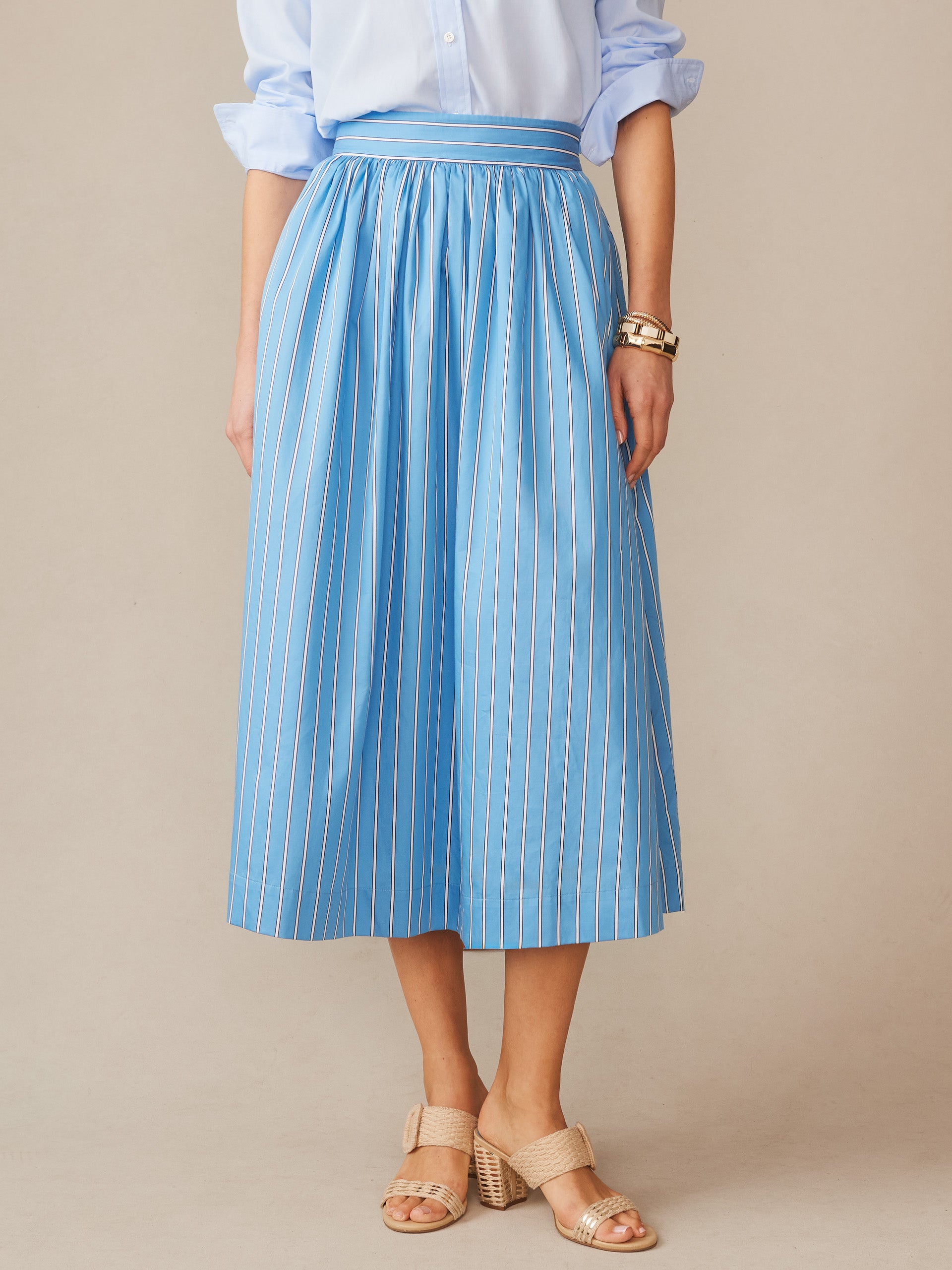Woman wearing a blue vertically striped Winnie Skirt in Stripe by J.McLaughlin paired with a light blue shirt and neutral heeled sandals, showcasing effortlessly stylish, enduring designs.