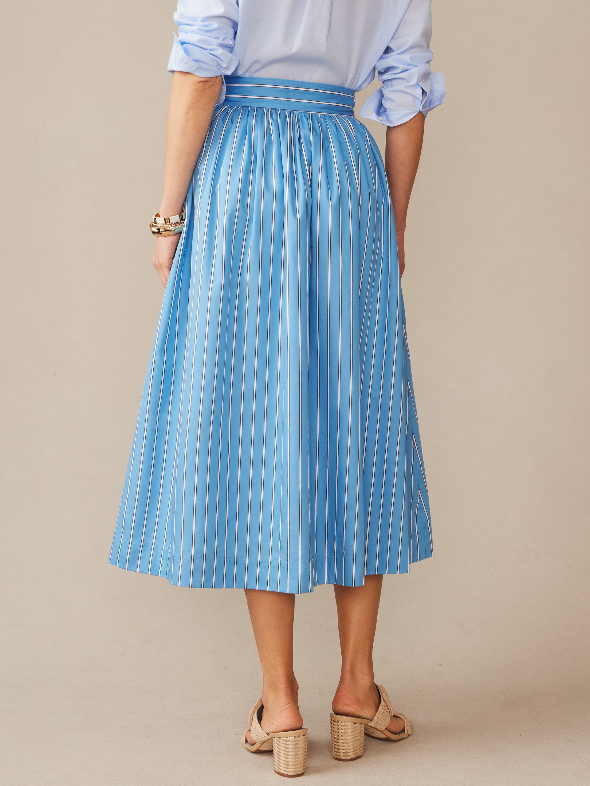 A woman wearing the Winnie Skirt in Stripe by J.McLaughlin, featuring a flowing blue and white striped pattern paired with a rolled-sleeve shirt and beige heels, effortlessly stylish with enduring designs and innovative fabrics, embodying a legacy of timeless fashion— a destination for defining style. Life, Well-Lived.