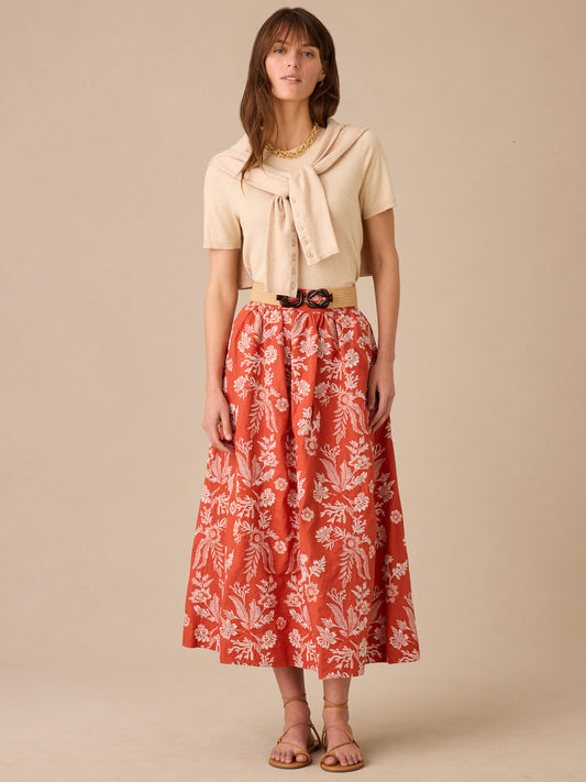 Woman wearing a beige top with a matching sweater draped over shoulders paired with a rust and off-white Winnie Linen Blend Embroidered Skirt by J.McLaughlin, styled with sandals and a woven belt, showcasing effortlessly stylish, enduring designs made from innovative fabrics.