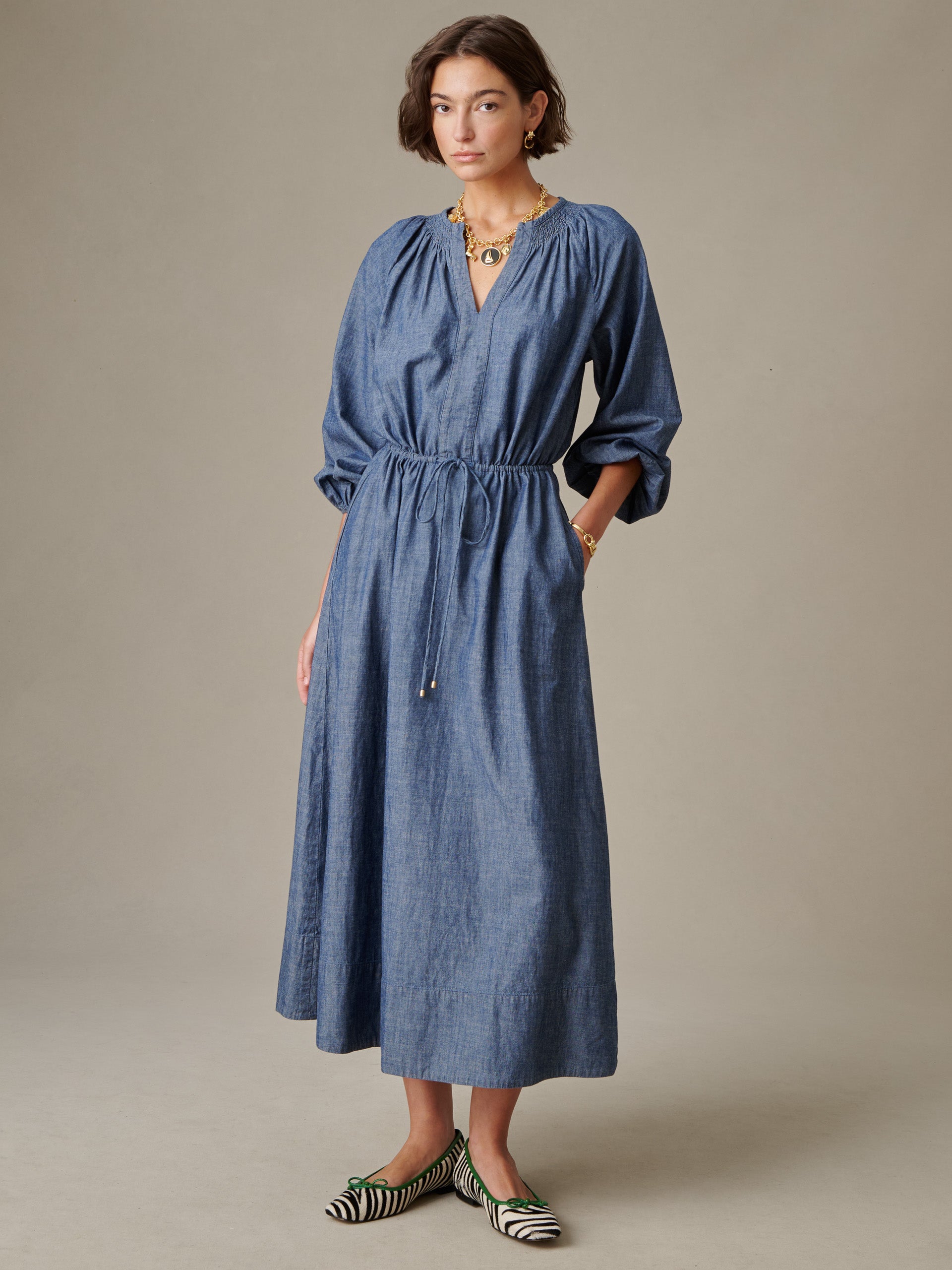 Woman wearing a blue Winslet Chambray Dress by J.McLaughlin with long sleeves and a drawstring waist, paired with bold black and white striped flats, showcasing effortlessly stylish, enduring design.