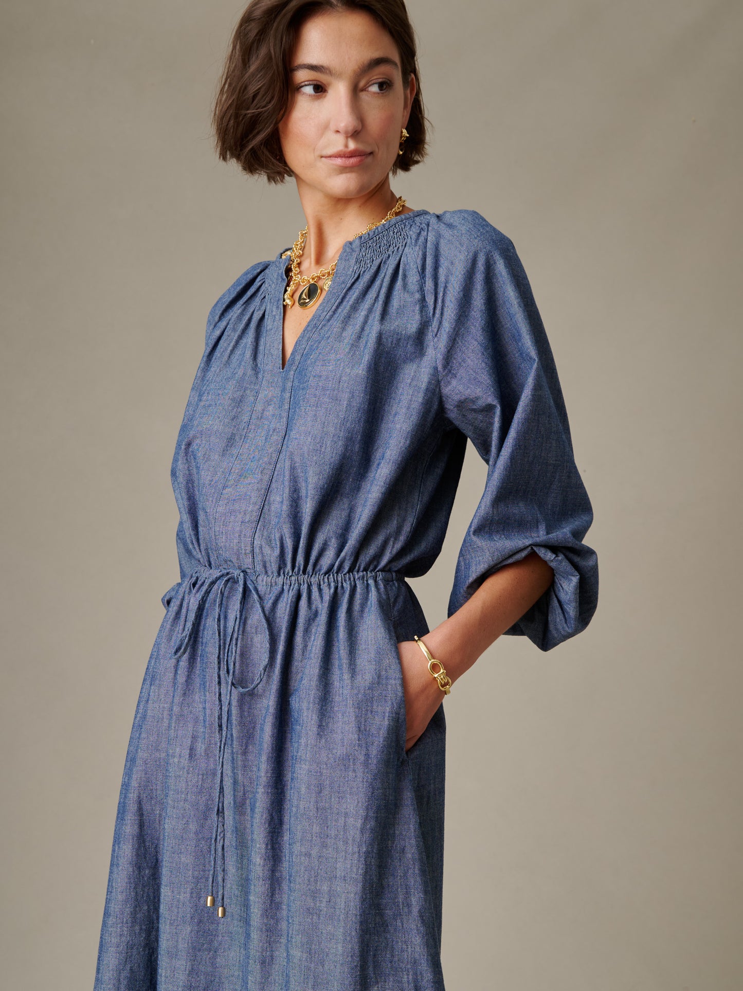 Woman wearing a blue Winslet Chambray Dress by J.McLaughlin featuring a gathered waist and three-quarter sleeves, styled with gold jewelry for an effortlessly stylish look.