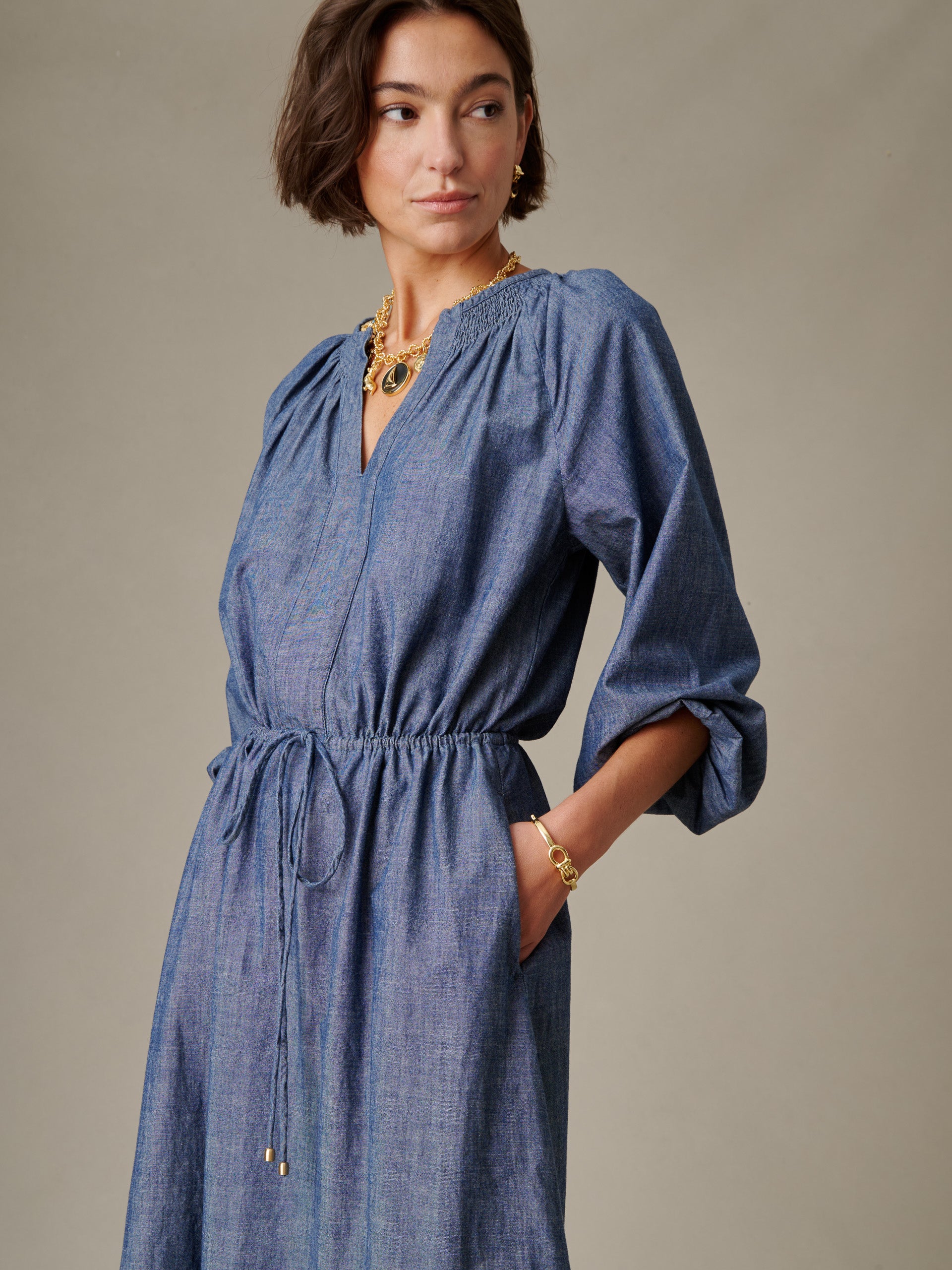 Woman wearing a blue Winslet Chambray Dress by J.McLaughlin featuring a gathered waist and three-quarter sleeves, styled with gold jewelry for an effortlessly stylish look.
