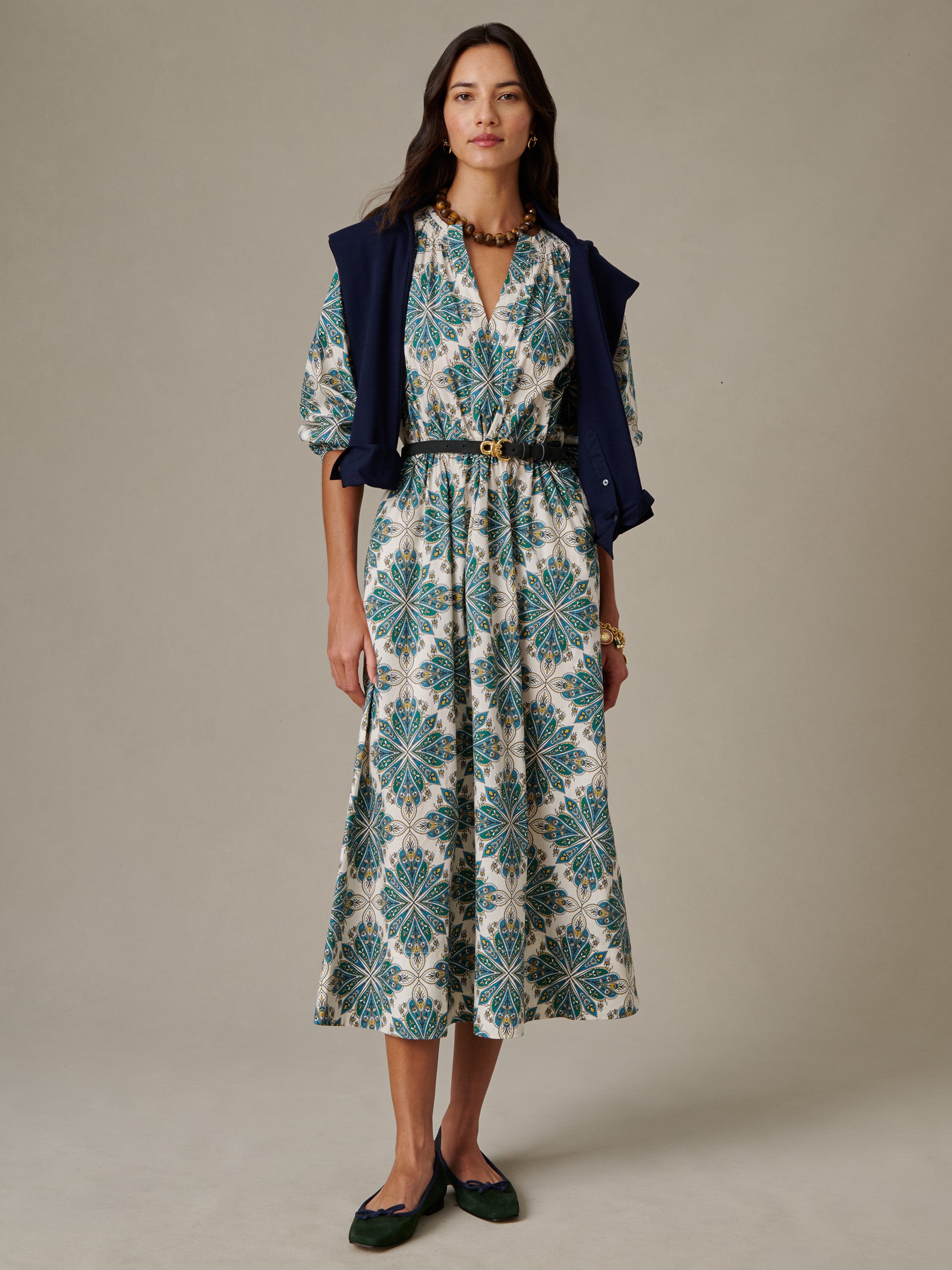 Woman wearing the Winslet Dress in Neo Radiant by J.McLaughlin, featuring a green and blue patterned midi dress with a black belt, paired with a navy sweater draped over her shoulders and dark flats.