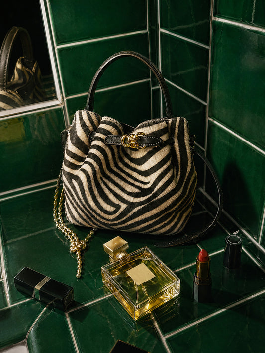 The Mini Lex Pony Hair Handbag in Zebra Stripes by J.McLaughlin displayed on green tiles with gold chain, perfume bottle, and red lipstick, showcasing timeless, stylish design.